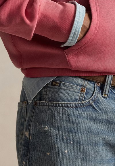 Χέρι σε κόκκινη μπλούζα με τσέπη πάνω από ανοιχτό μπλε τζιν τζιν με πιτσιλιές μπογιάς, στρωμένο με μπλε πουκάμισο από κάτω.