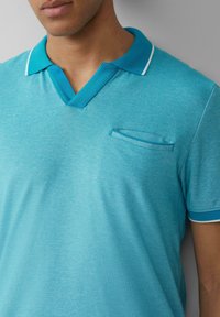 Polo turquoise avec un col en V, un col contrastant et une petite poche poitrine. Le tissu a un aspect texturé avec des accents blancs.