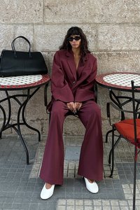 Bordo oversize blejzer in ujemajoče hlače, bele ravne čevlje, sončnih očal. Črna torbica leži na okrogli mizi s pisanem vrhom.