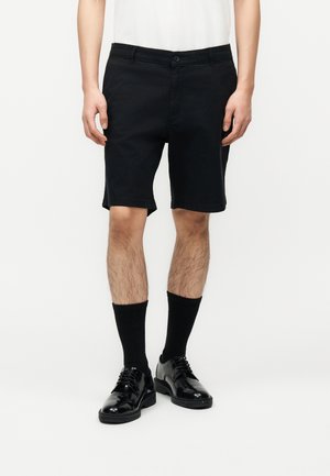 Homme portant un short noir jusqu'aux genoux, des chaussettes noires et des chaussures habillées noires brillantes, debout devant un fond blanc.