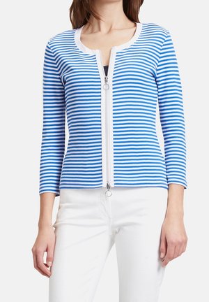 Frau trägt eine blau-weiß gestreifte Langarm-Strickjacke mit Reißverschluss, kombiniert mit weißen Hosen, abgebildet von Hals bis Hüfte.