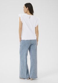 Haut blanc à manches courtes avec bord en dentelle et ouverture en goutte d'eau à l'arrière, associé à un jean bleu clair à jambes larges. Texture de tissu lisse.