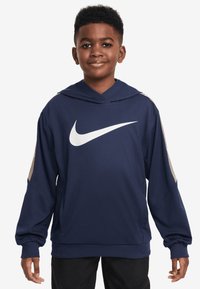 Zēns smejas, ģērbts tumši zilā Nike kapucē ar baltu Swoosh logotipu, stāv ar rokām atpūstās pozās uz balta fona.