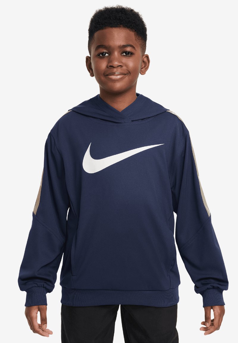 Zēns smejas, ģērbts tumši zilā Nike kapucē ar baltu Swoosh logotipu, stāv ar rokām atpūstās pozās uz balta fona.