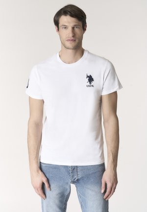 Uomo in piedi, di fronte, che indossa una maglietta bianca con il logo USPA e jeans azzurri chiari, davanti a uno sfondo chiaro e uniforme.