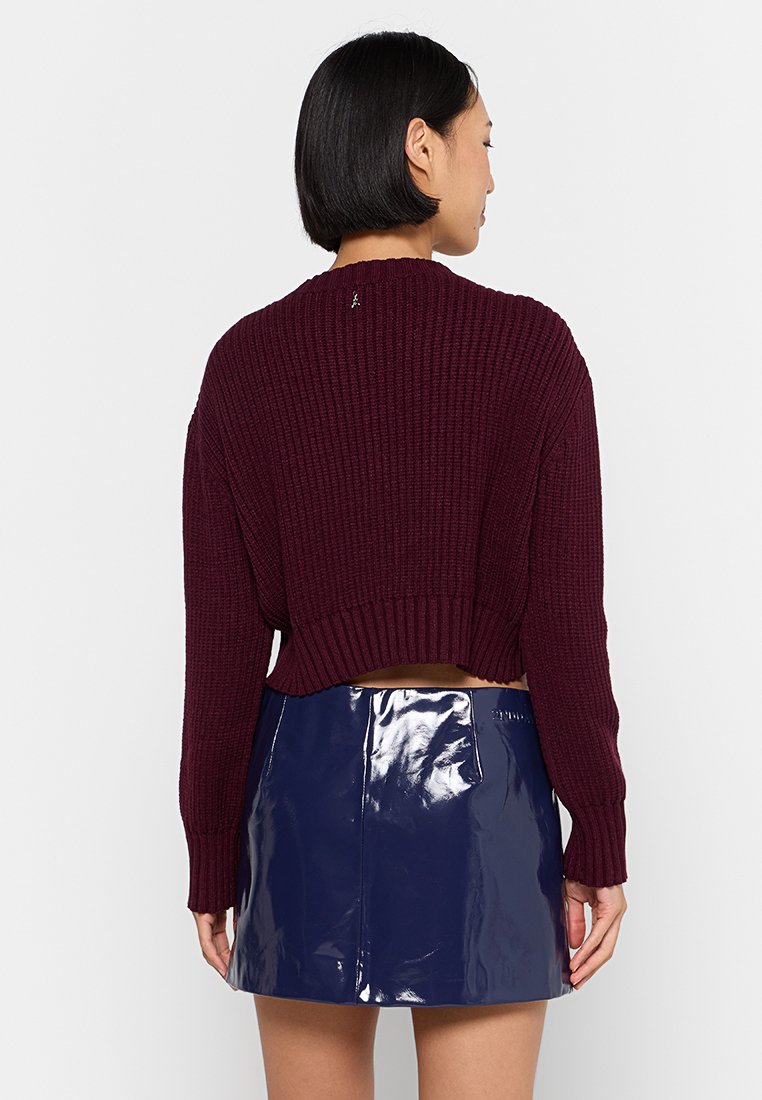 Donna con i capelli neri corti che indossa un maglione lavorato a maglia bordeaux e una mini gonna blu navy lucida, rivolta di spalle su uno sfondo bianco.