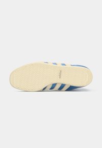 Blue athletic shoe with a textured rubber sole, featuring three beige stripes and a curved shape. The surface displays a smooth finish.