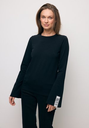 Vrouw met recht lichtbruin haar, gekleed in een zwart shirt met lange mouwen met uitlopende mouwen en witte tekst op één manchet, voor een effen achtergrond.