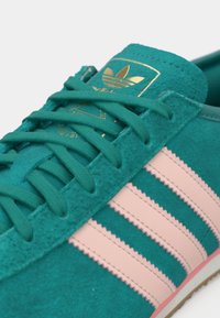 Τιρκουάζ δερμάτινο αθλητικό παπούτσι με ροζ ρίγες, που διαθέτει επίπεδα κορδόνια και χρυσή ετικέτα λογότυπου στη γλώσσα. Ομαλή υφή με πλευρικές διακοσμήσεις.