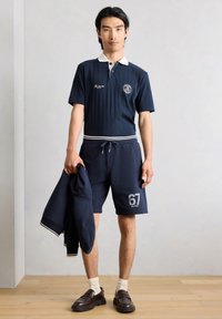 Άνδρας φοράει μπλε polo μπλούζα και σορτς με αριθμό 67, κρατάει ταιριαστό μπουφάν, στέκεται σε ξύλινο πάτωμα μπροστά σε απλό φωτεινό τοίχο.