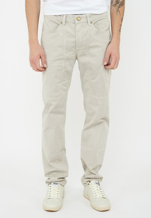 Pantaloni beige a gamba dritta con tasche applicate anteriori, indossati da una persona con sneakers bianco sporco e una maglietta bianca, su uno sfondo bianco semplice.