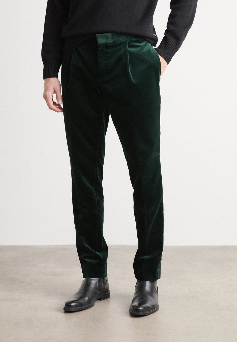 Dark green velvet trousers with a smooth texture, featuring pleats and side pockets, paired with black leather ankle boots.