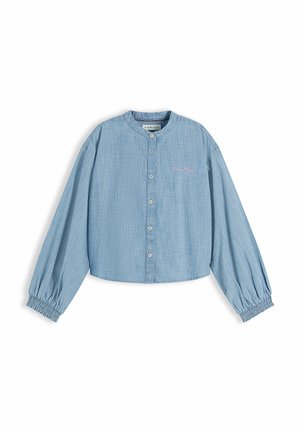 Niña joven de pie con una camisa azul abotonada, falda plisada beige y mocasines negros sobre un fondo blanco liso.