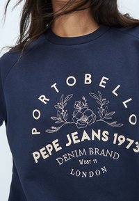 Námorně modrá mikina s dekorativní květinovou výšivkou. Tisk s textem "Pepe Jeans 1973" a "Portobello" v světlé béžové barvě. Kulatý výstřih, raglánové rukávy.