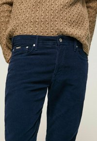 Person som bär en brun kabelstickad tröja instoppad i marinblå kordflortbyxor med synlig knapp och bälteshällor.