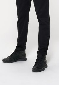 Czarne wysokie sneakersy mają połączenie gładkiej i strukturalnej skóry, wyraźne sznurowanie oraz wyściółkę w kołnierzu, zestawione z dopasowanymi czarnymi spodniami.