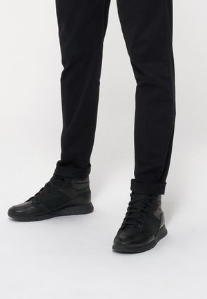 Czarne wysokie sneakersy mają połączenie gładkiej i strukturalnej skóry, wyraźne sznurowanie oraz wyściółkę w kołnierzu, zestawione z dopasowanymi czarnymi spodniami.