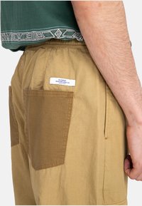Homme portant un short kaki avec de grandes poches arrière et une chemise verte avec un ourlet à motifs, se tenant avec un bras détendu à ses côtés.