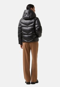 Zwarte pufferjack met een grote capuchon, doorgestikt ontwerp en glanzende textuur. Gepaarde met bruine wijde broek en zwarte schoenen.