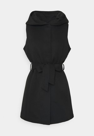 Veste noire sans manches ceinturée avec un large col, une taille cintrée et un ourlet arrivant aux genoux sur fond blanc.