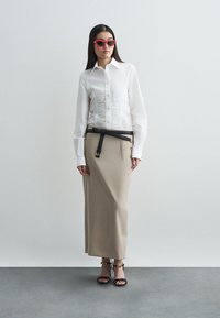 Camisa blanca de botones con delantero texturizado, falda beige de talle alto, cinturón negro y gafas de sol rojas. Las sandalias tienen un diseño de correas.