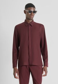 Camisa burgundy de manga larga hecha de tejido ligero, con cierre de botones en el frente, cuello con puntas y corte recto, acompañada de gafas de sol.
