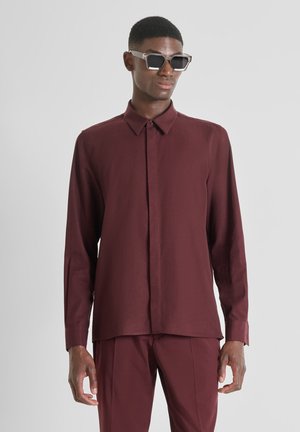 Camisa burgundy de manga larga hecha de tejido ligero, con cierre de botones en el frente, cuello con puntas y corte recto, acompañada de gafas de sol.