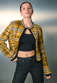 Cárdigan de punto a cuadros en amarillo y negro con ribete negro y collar adornado, llevado sobre un top corto negro y jeans negros ajustados.