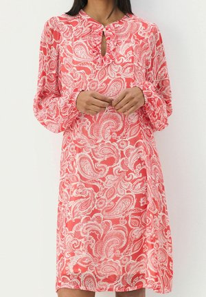 Frau trägt ein korallenfarbenes, langärmliges Kleid mit weißen Paisley-Mustern und einem kleinen, geknöpften Schlüssellochausschnitt, Hände auf der Taille verschränkt.