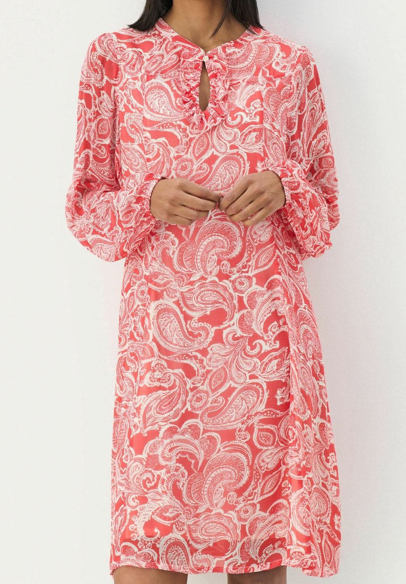 Femme portant une robe corail à manches longues avec des motifs cachemire blancs et un petit décolleté en forme de trou de serrure boutonné, les mains jointes à la taille.