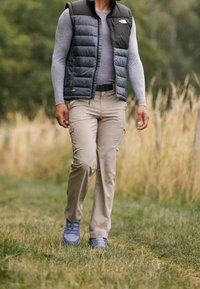 Schwarz-graue isolierte Weste über einem grauen Langarmshirt, beigefarbene Cargohosen und blaue Turnschuhe, die auf einem grasbewachsenen Weg gehen.