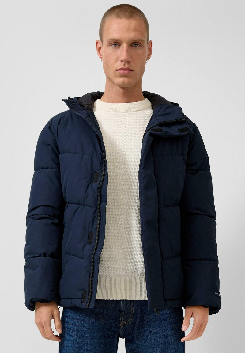 Navy pufferjack met rits, een vaste capuchon en gestructureerde manchetten. G gedragen over een crèmekleurige gebreide trui en blauwe jeans.