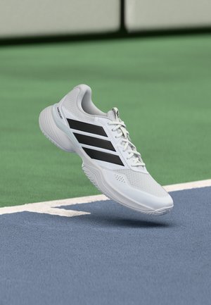 Chaussure de sport blanche avec des bandes noires flottant au-dessus d'une surface de court de tennis bleue et verte près d'une ligne blanche.