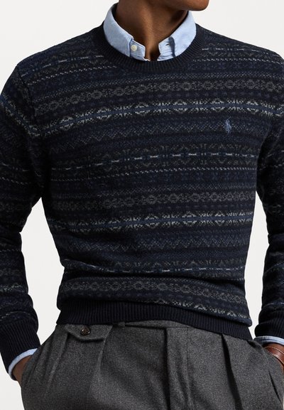Navy patterned sweater with a ribbed crew neckline, featuring various geometric designs in shades of blue and gray, paired with a light blue shirt.