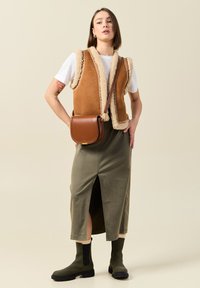 Beige Shearling-Weste mit brauner Handtasche, olivgrüner Midirock mit seitlichem Schlitz und schwarzen Stiefeletten mit elastischen Seiten.