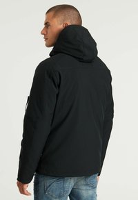 Chaqueta negra con capucha y acabado suave, que cuenta con un bolsillo con cremallera en el brazo izquierdo y un corte recto. Combinada con jeans azules.