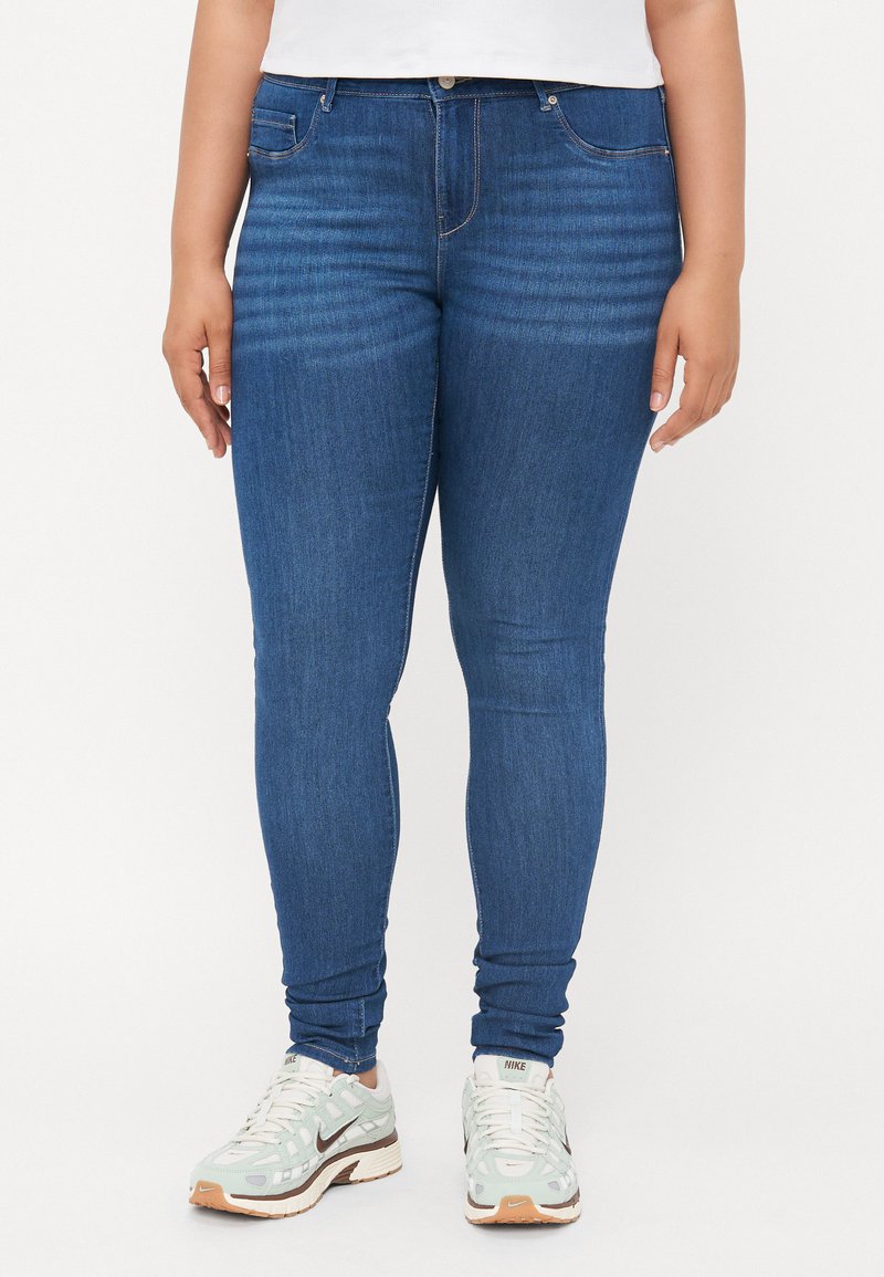 Jeans ajustados azules de mezclilla, con un diseño clásico de cinco bolsillos, costuras claras y una silueta ajustada, combinados con zapatillas blancas.