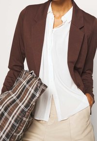 Brun lättviktsblazer med krage, bärs över en vit skjorta med knappar. Håller en handväska med rutigt mönster i dämpade färger.