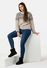 Maglione beige con scollo decorato, jeans blu e stivaletti neri alla caviglia. Seduto in posa su blocchi bianchi contro uno sfondo semplice.