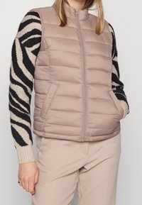 Vesta s límcem a vycpávkami v tlumené béžové barvě, s předním zipem a bočními kapsami. Nositelná přes černý a krémově pruhovaný svetr s dlouhým rukávem.