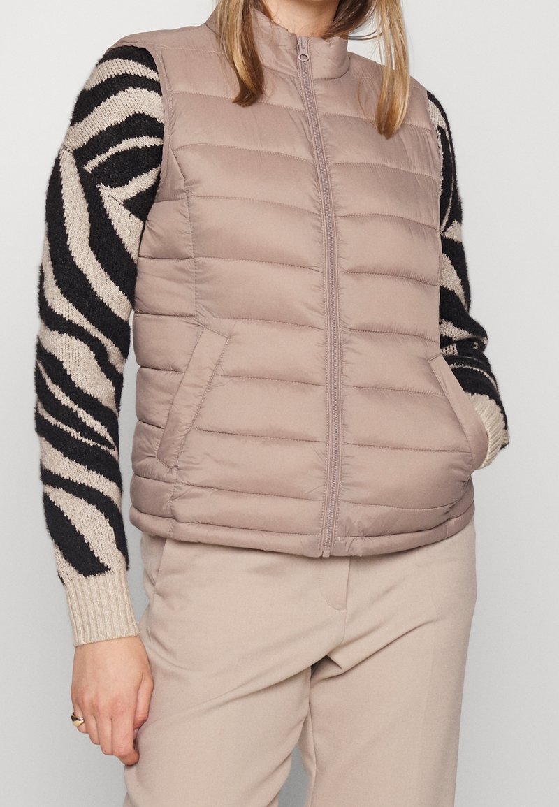 Vesta s límcem a vycpávkami v tlumené béžové barvě, s předním zipem a bočními kapsami. Nositelná přes černý a krémově pruhovaný svetr s dlouhým rukávem.