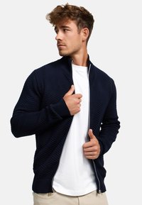 Marineblå strikkjakke med fiskebensmønster, hel glidelås, stående krage, ribbestrikkede mansjetter, båret over en hvit t-skjorte, teksturert stoff.