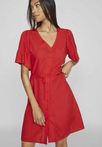 Abito rosso con bottoni, scollatura a V, maniche corte a sbuffo, cintura legata in vita e gonna svasata. Tessuto dalla texture liscia.