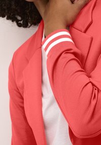Koralowa marynarka z teksturowanym wykończeniem, z kontrastowymi mankietami w białe paski. Projekt obejmuje dopasowaną sylwetkę i kanciaste klapy.