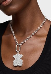 Collier en chaîne en argent avec un pendentif en forme d'ours à la surface texturée, posé sur un haut noir et une peau lisse.