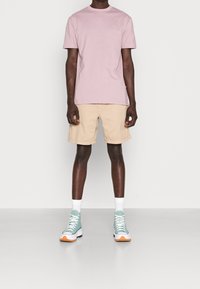 Camiseta de algodón rosa, pantalones cortos beige y zapatillas altas en color teal con detalles en blanco. El modelo está de pie contra un fondo liso.