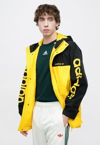 Μαύρο και κίτρινο αδιάβροχο με λογότυπο «adidas». Διαθέτει κουκούλα, πλαϊνές τσέπες και σχεδιασμό με αντίθετα χρώματα. Το μοντέλο φοράει πράσινο πουκάμισο και λευκό παντελόνι.