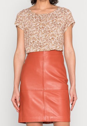 Frau trägt eine kurzärmelige, geblümte Bluse, in einen hoch taillierten, rostfarbenen Lederrock gesteckt.