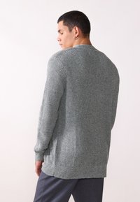 Grå stickad tröja med ett texturerat mönster, rund halsringning och ribbade ärmslut och nederkant. Bärs över mörka byxor.