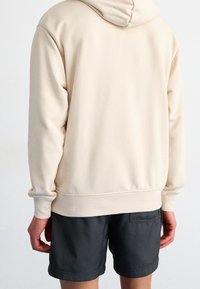Beige hooded sweatshirt with long sleeves, soft texture, and a ribbed hem, paired with dark gray shorts.